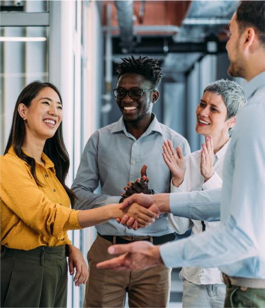 Des collègues se serrent la main dans un bureau tandis que d’autres applaudissent, symbolisant une réussite collective.