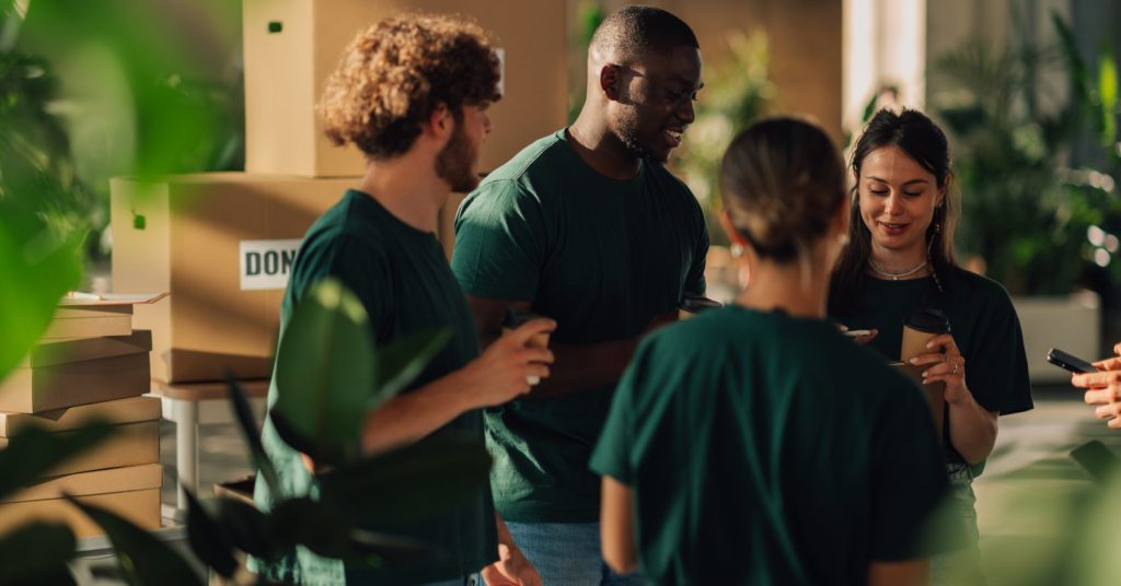 Groupe de bénévoles portant des t-shirts verts discutant autour de cartons empilés dans un espace un intérieur lumineux.